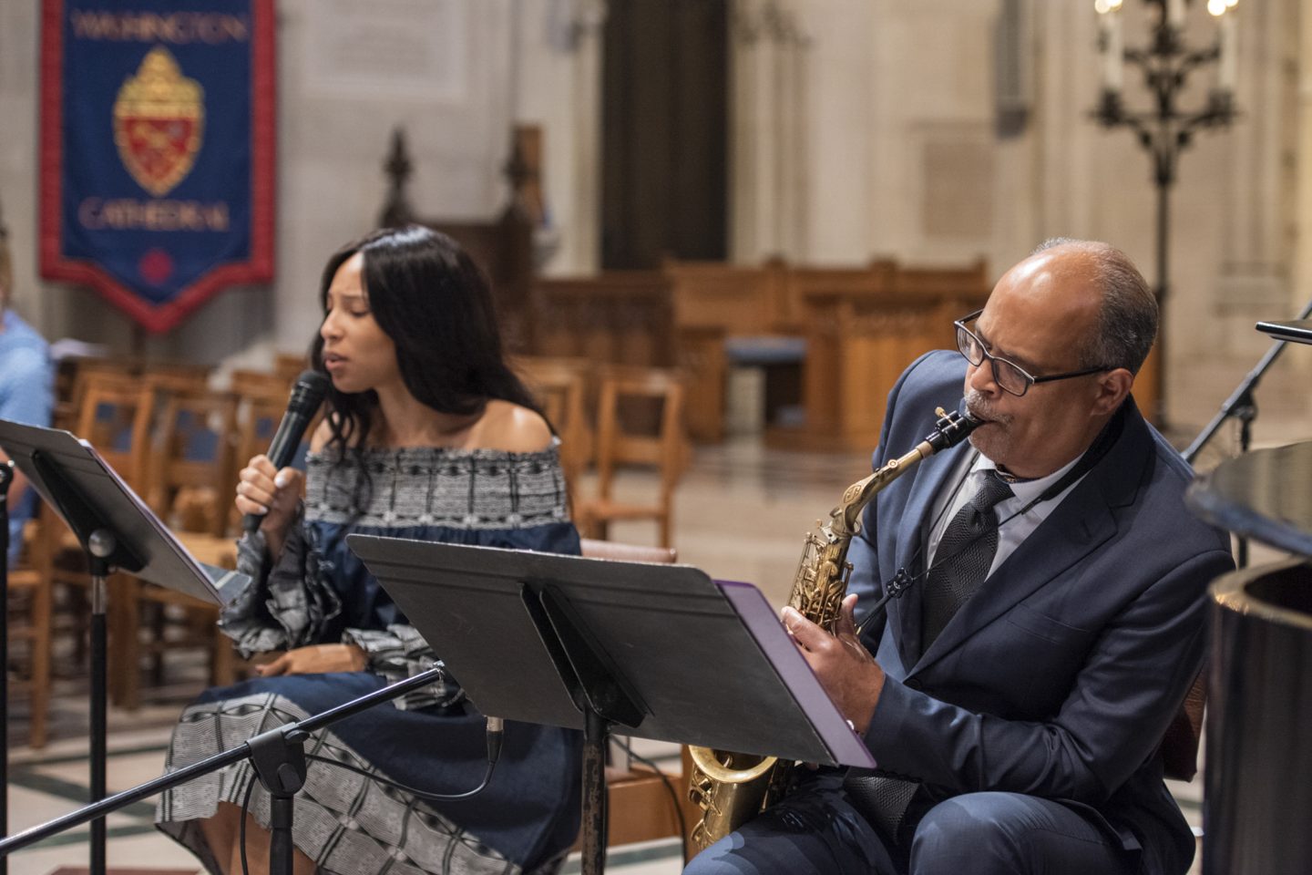 Singer singing and holding a microphone next to a saxophonist playing the saxophone, both seated, in the Cathedral