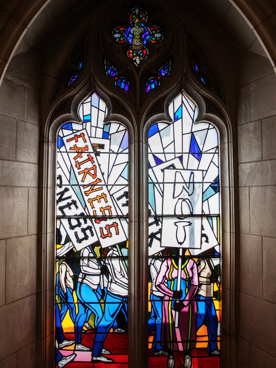 Two stained-glass windows depicting people walking and standing with protest signs that read “Fairness” and “Not”