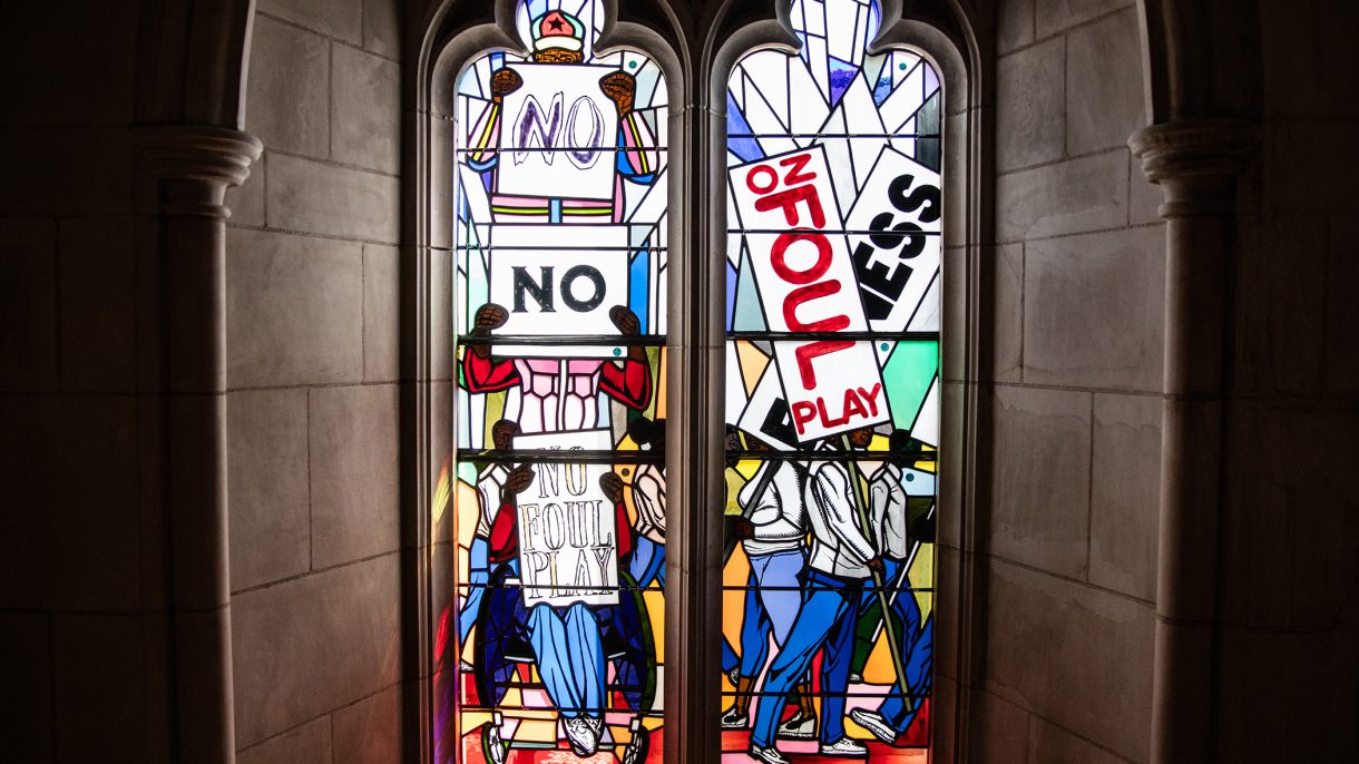 Two stained-glass windows depicting people walking and standing with protest signs that read “NO” and “No Foul Play"”