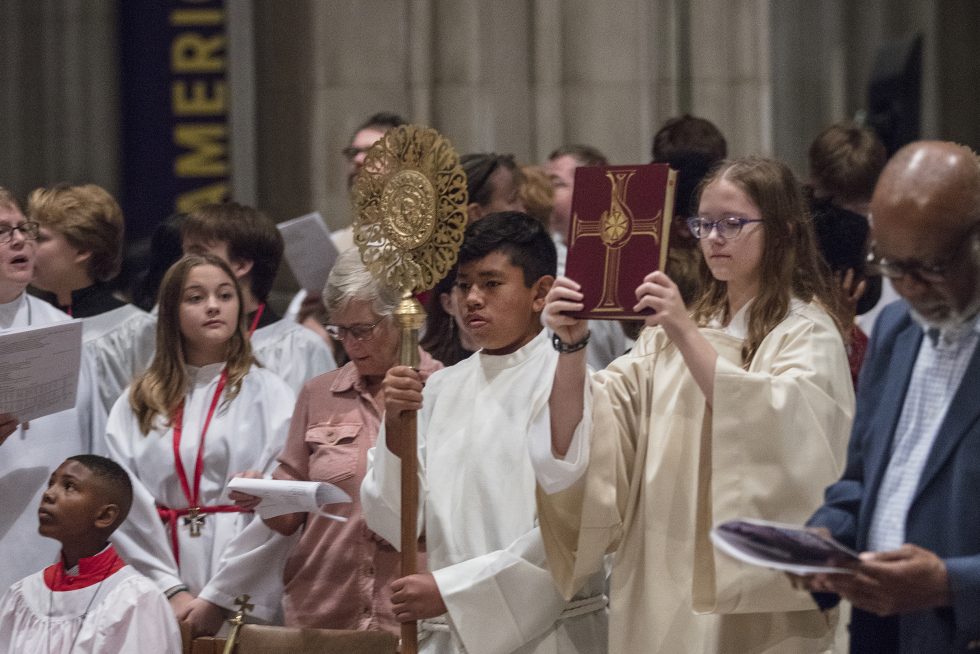 Two children in acolyte vestments carry a bible and a processional staff. Other service attendees watch in the background.