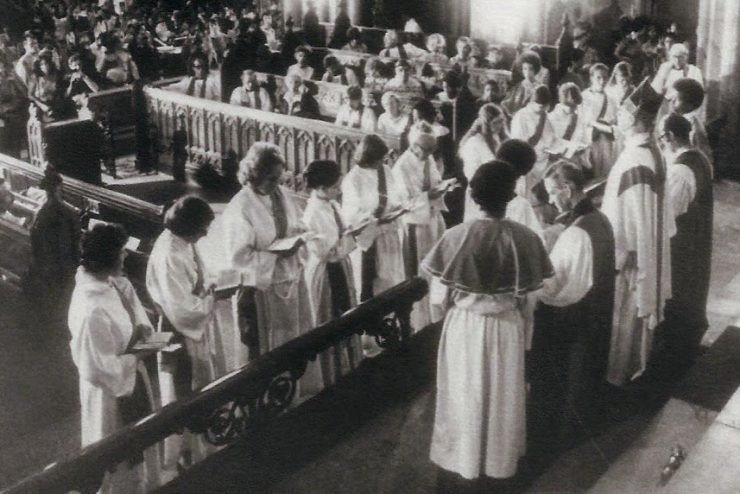 Eleven women wearing deacon robes stand in a church before clergy holding books and surrounded by pews and congregants.