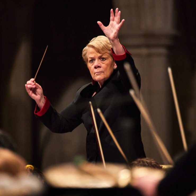 Conductor Marin Alsop dressed in black top holds baton with arm raised in front of orchestra.