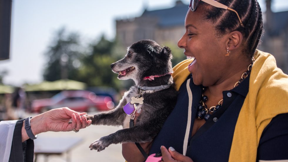 A clergy person's hand shakes a dog's paw in greeting, and the woman holding the dog laughs.