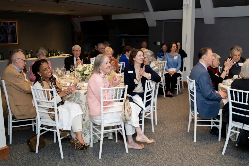 Groups of adults applaud while seated at round tables.